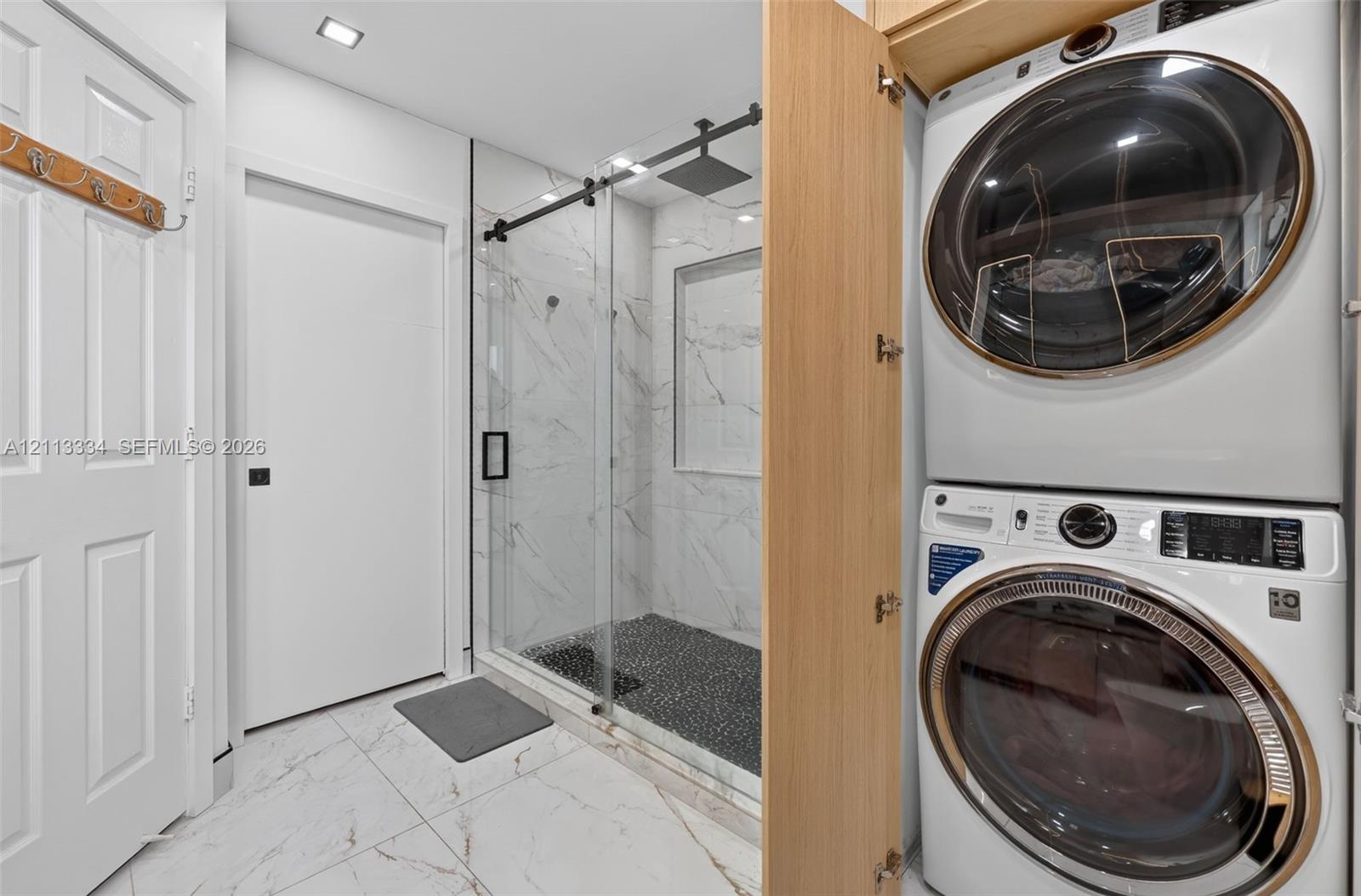Walk-in shower with stone-look wall tile, recessed shelving, a glass sliding door with matte black hardware, and a pebble-tiled floor, featuring built-in stacked laundry appliances behind cabinetry