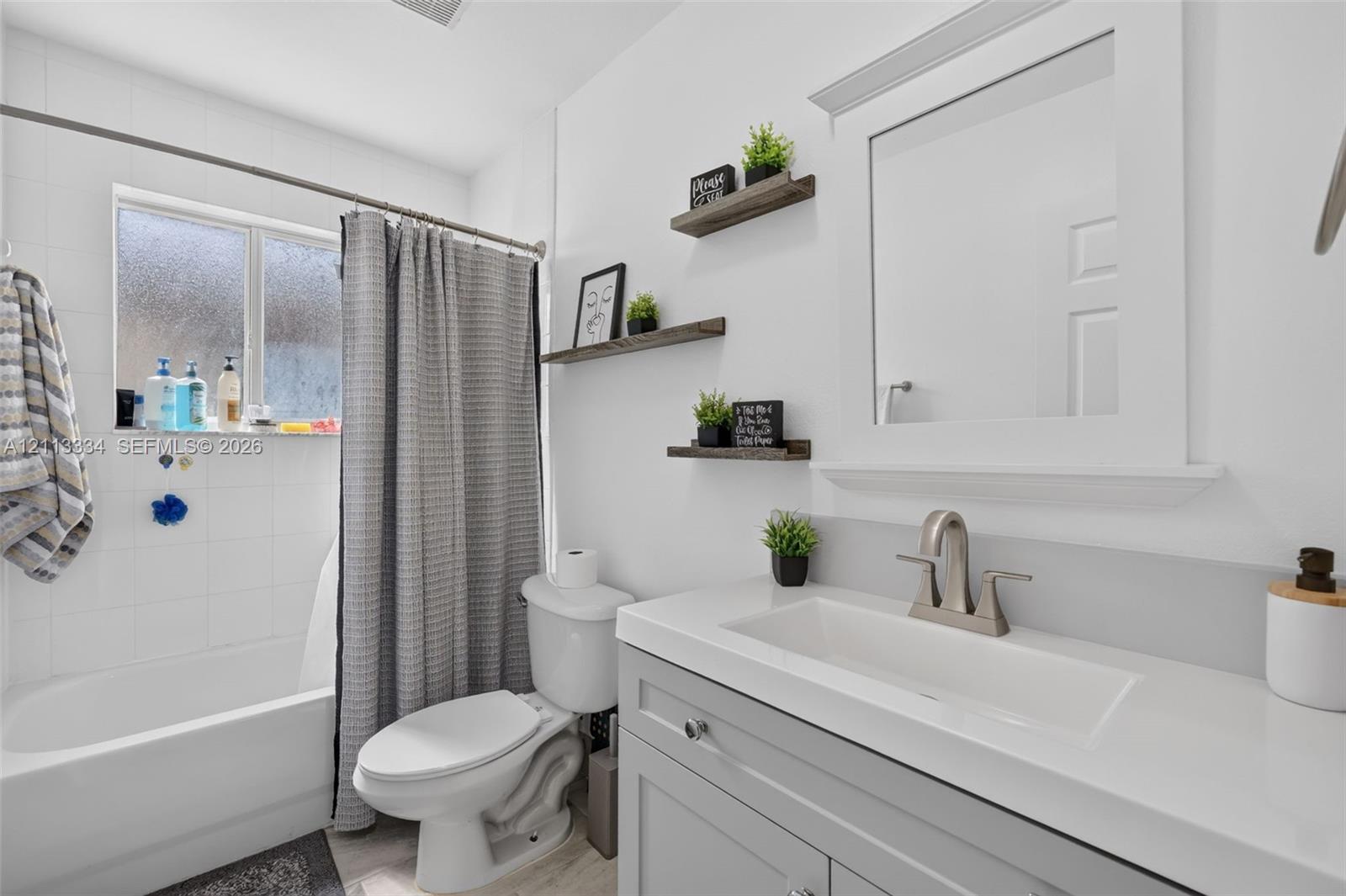 Bathroom featuring a gray vanity with an integrated rectangular sink, brushed nickel faucet, white-tiled shower surround, and three wood-finish wall-mounted shelves