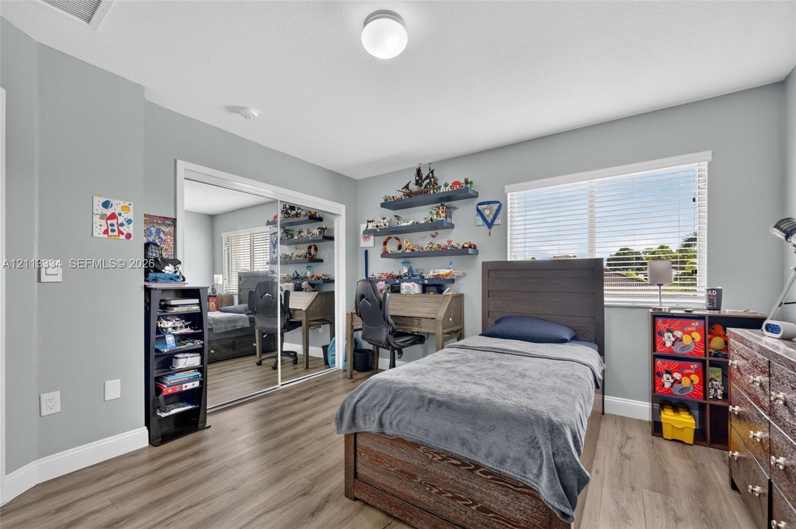 Wood-finish flooring, sliding mirror-panel closet doors, horizontal window blinds, and overhead flush-mount lighting