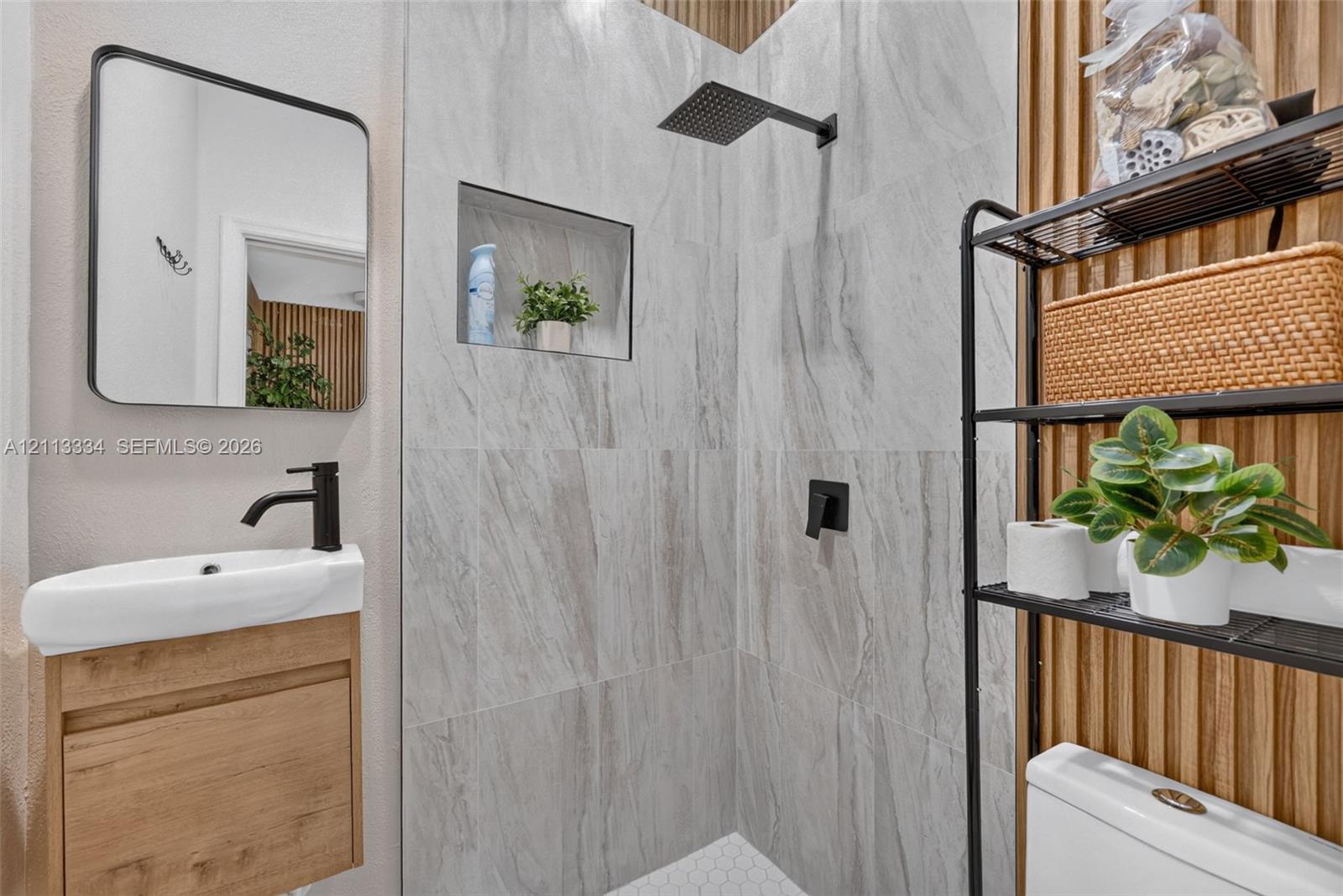 Wall-mounted wood-finish vanity with a rectangular sink and matte black faucet, wall-mounted mirror with black frame, walk-in shower featuring floor-to-ceiling stone-style tile, built-in shower niche, and wood-slat accent wall