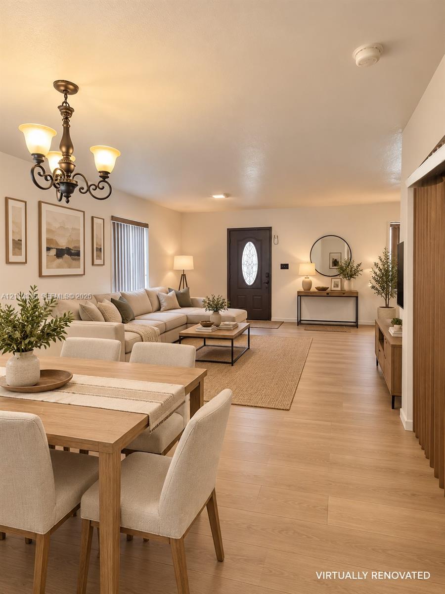 Open-concept interior featuring light wood-finish flooring, a decorative chandelier with classic scrolled arms, and a dark-toned exterior entry door with an oval glass insert Open-concept interior featuring light wood-finish flooring, a decorative chandelier with classic scrolled arms, and a dark-toned exterior entry door with an oval glass insert