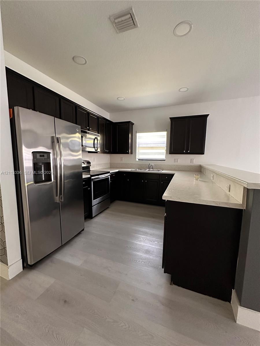 Kitchen featuring dark-finish cabinetry, light-toned stone-style countertops, stainless steel appliances, and wood-finish flooring
