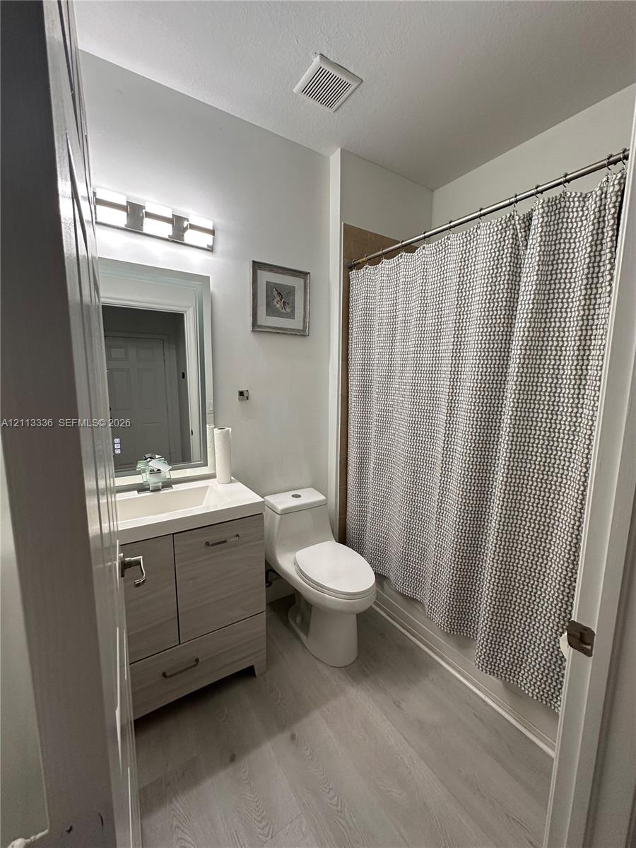 Bathroom featuring a wood-finish vanity with a rectangular sink, wall-mounted vanity lighting, a one-piece toilet, and wood-finish flooring