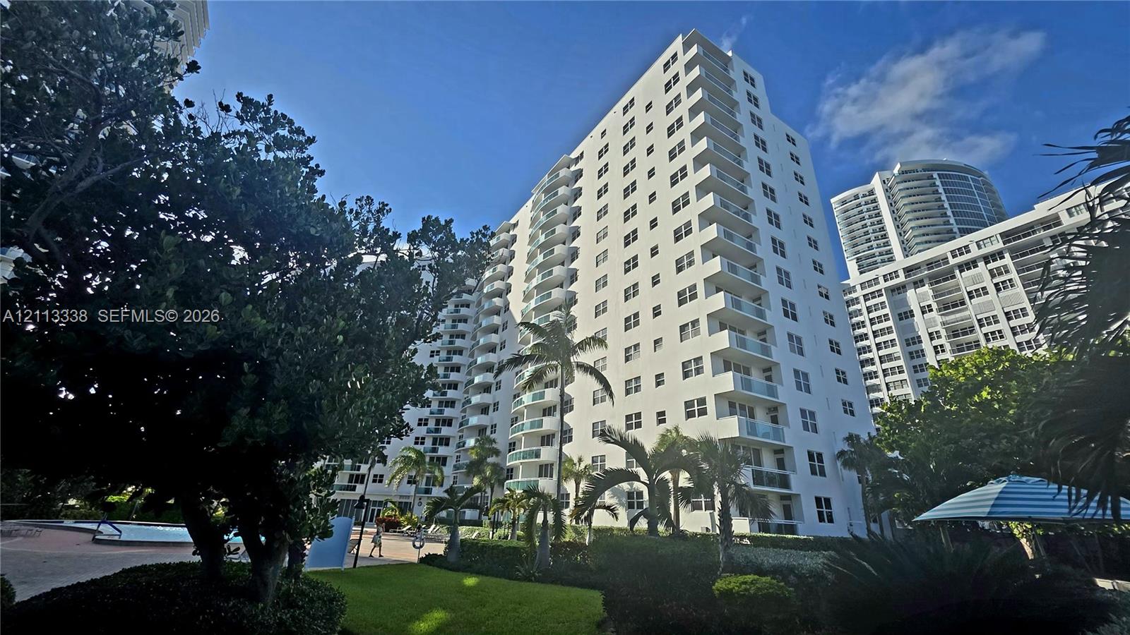 High-rise residential building featuring white stucco facade, stacked private balconies with glass railings, surrounding mature landscaping, and an outdoor swimming pool