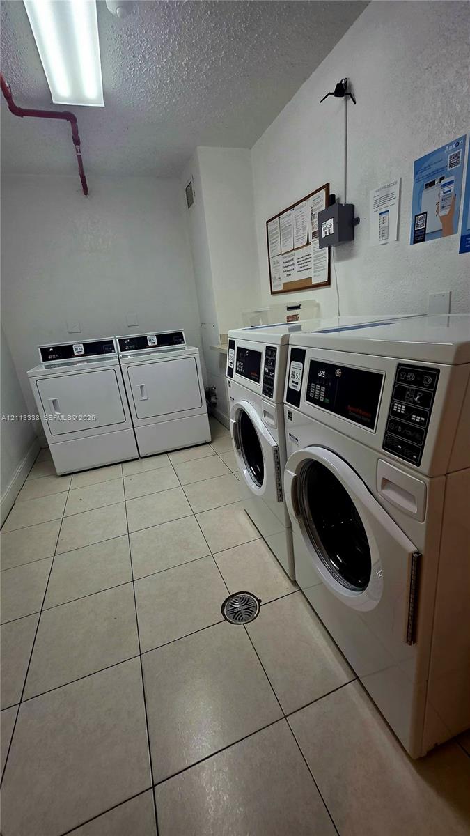 Laundry facility equipped with four commercial-grade front-load washers and dryers, white-toned ceramic tile flooring, and integrated ceiling lighting