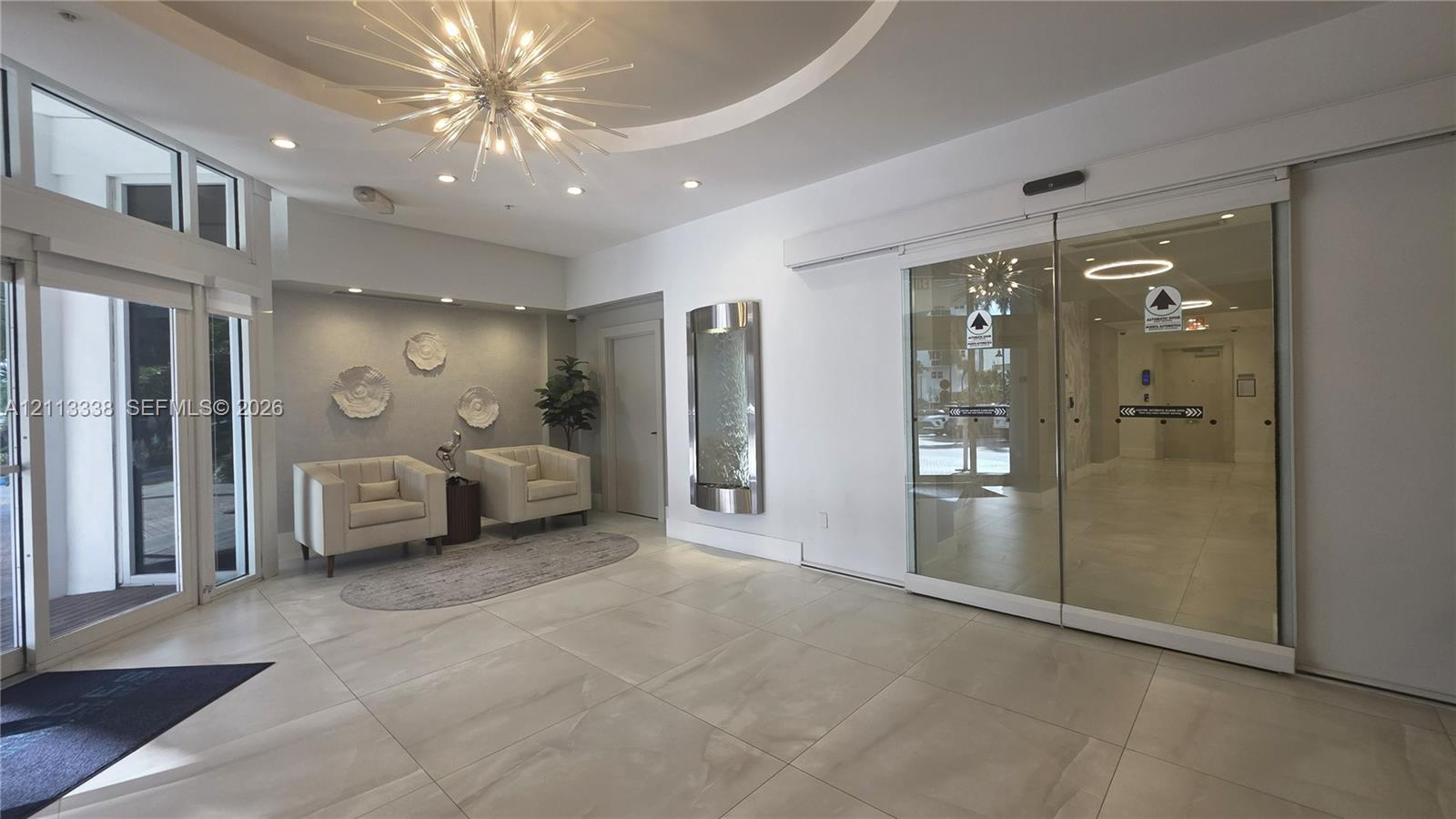 Entrance lobby featuring large-format tile flooring, a recessed circular ceiling detail, a geometric starburst chandelier, integrated recessed lighting, and a wall-mounted metal water feature