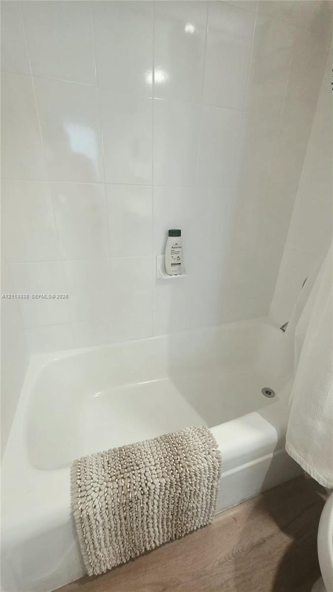 Bathtub with integrated shower, white wall tile surround, built-in soap niche, and wood-finish flooring