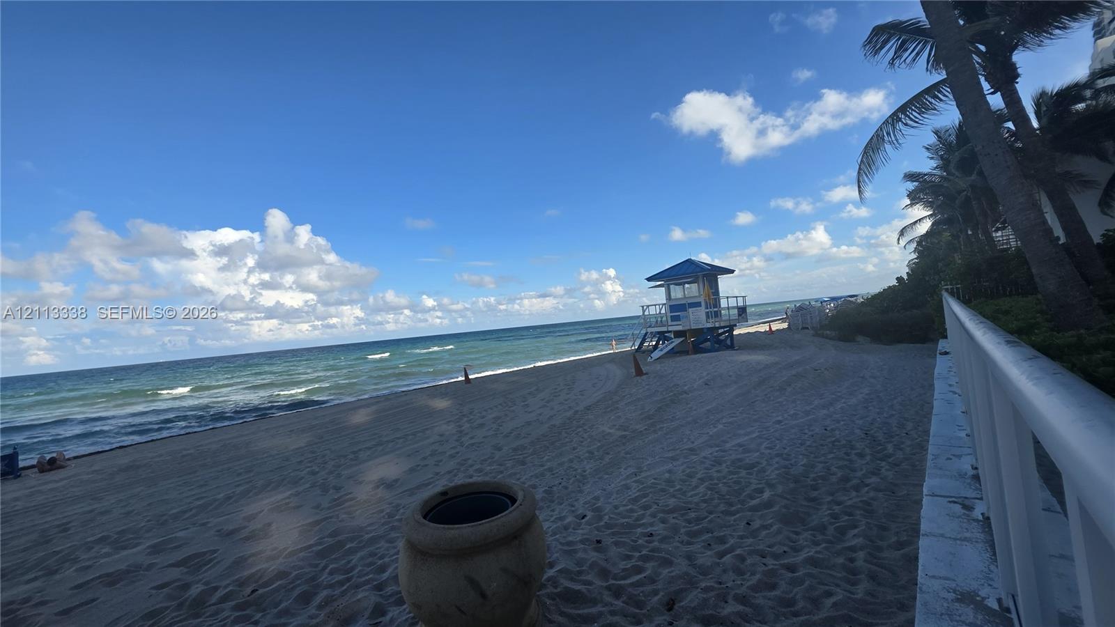 Sandy beachfront property featuring a white metal perimeter railing, mature palm trees, and coastal ocean scenery