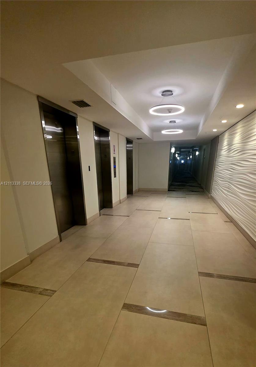 Elevator lobby featuring metallic-finish doors, large-format tile flooring with inlaid stone accents, textured three-dimensional wall paneling, and circular LED pendant lighting