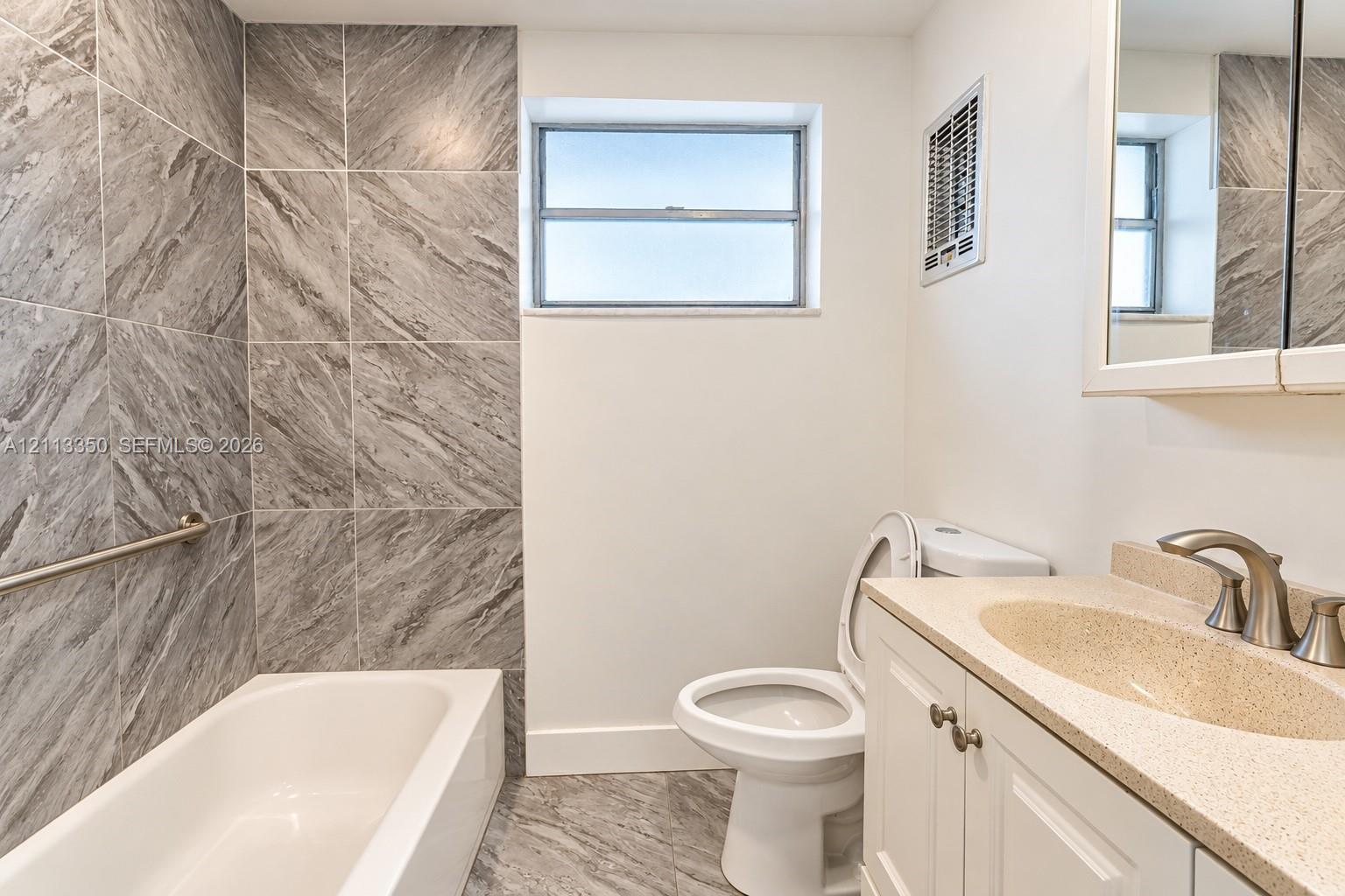 Bathroom featuring a tub-shower combination with large-format grey tile surround, stone-finish countertop, white cabinetry with metallic hardware, and a medicine cabinet with an integrated mirror