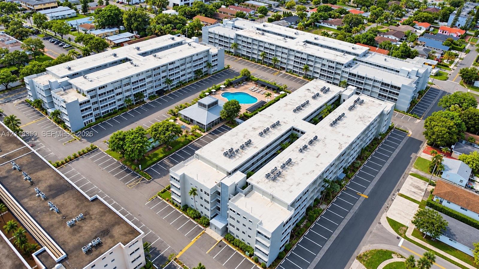 Aerial perspective of a multi-story residential complex featuring paved surface parking, an outdoor swimming pool, and a dedicated clubhouse structure