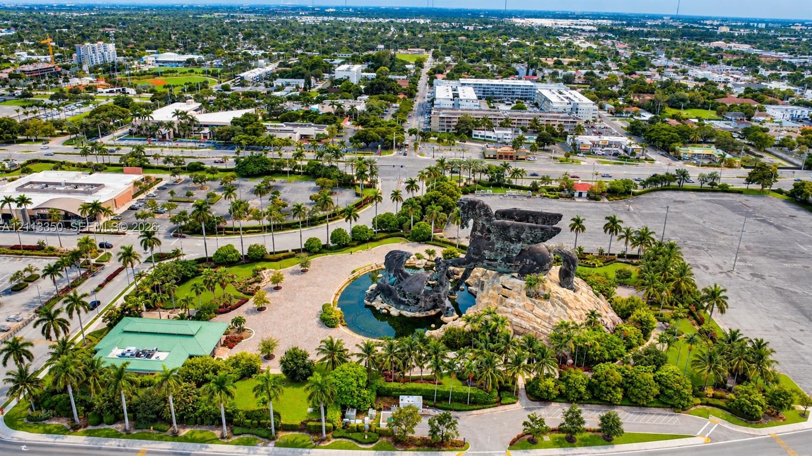 Aerial perspective featuring a large-scale sculptural monument, central water feature, paved courtyards, and extensive mature palm tree landscaping