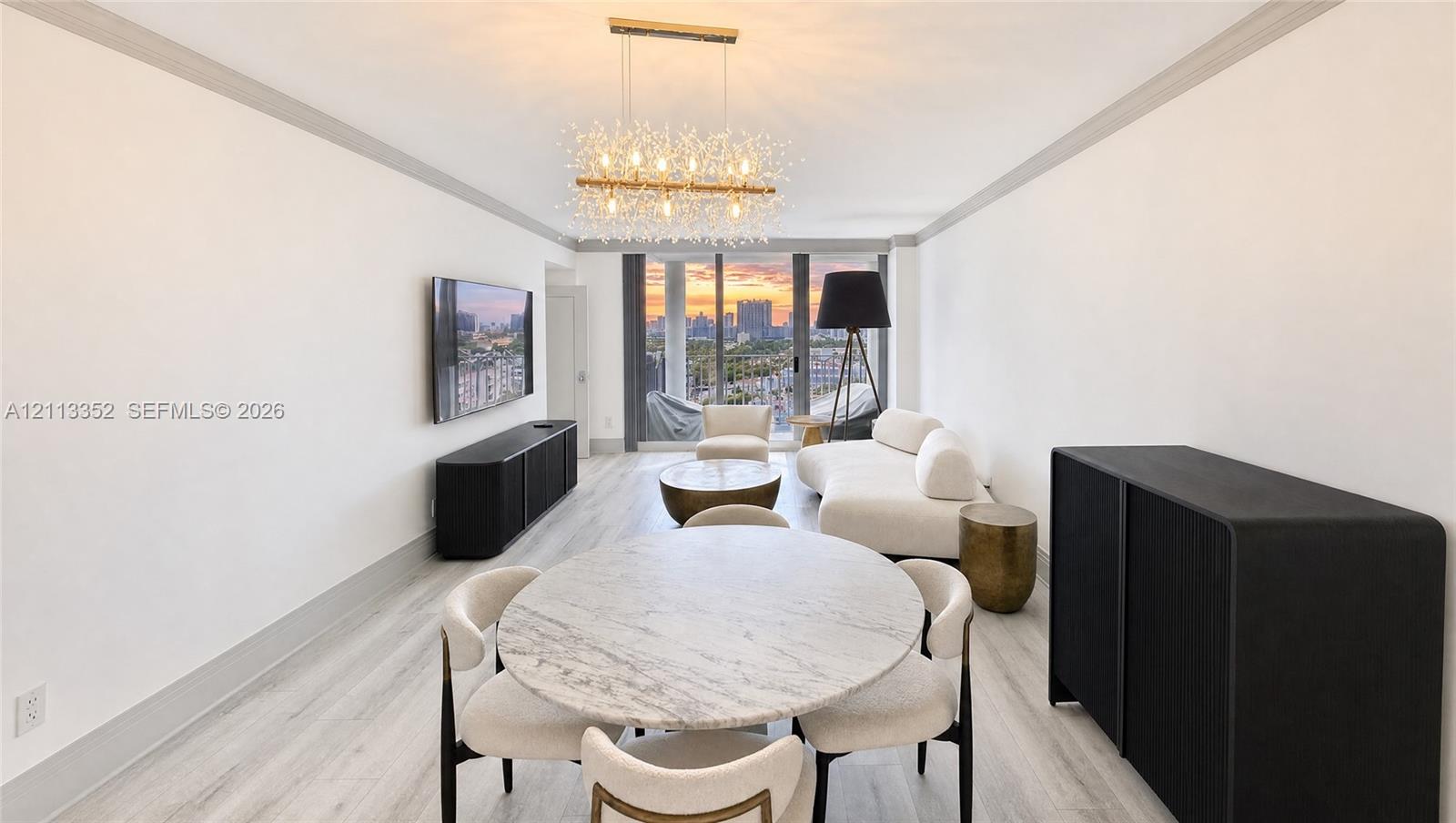 Open-concept area featuring light-toned wood-finish flooring, a multi-bulb chandelier, crown molding, and floor-to-ceiling glass doors with an urban sunset backdrop