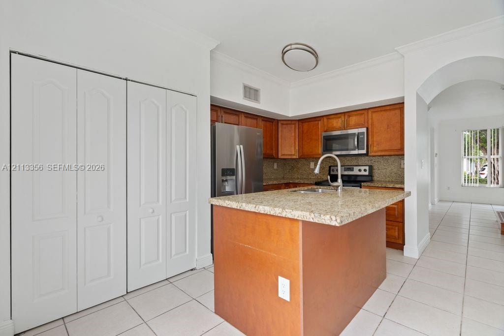 Kitchen featuring wood cabinetry, stone countertops, stainless steel appliances, a center island with an integrated sink, and tile flooring