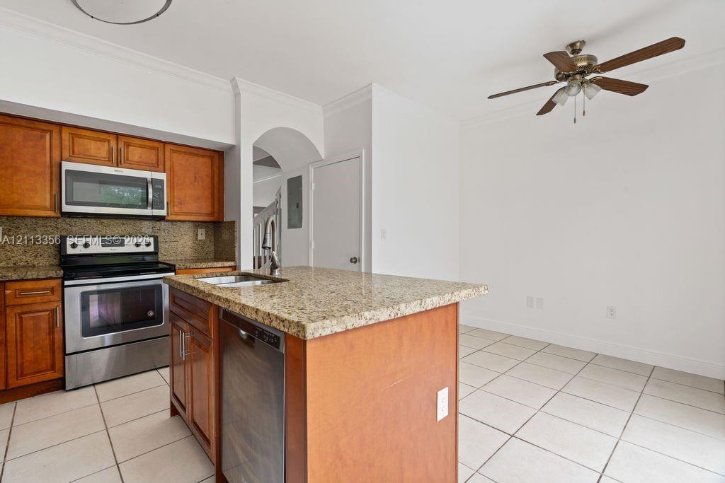 Kitchen featuring warm-tone wooden cabinetry, stone countertops, stainless steel appliances, a center island with a built-in dishwasher, and ceramic tile flooring