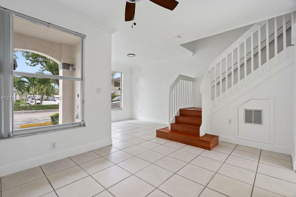Open-concept interior featuring light-toned tile flooring, large windows, a ceiling fan, and a staircase with wood-finish treads and white balusters