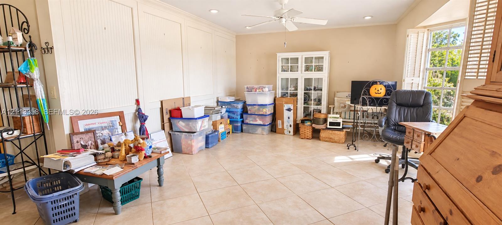 Room interior featuring tiled flooring, wall paneling, a ceiling fan with integrated lighting, built-in display cabinetry, and multi-pane windows with plantation-style shutters