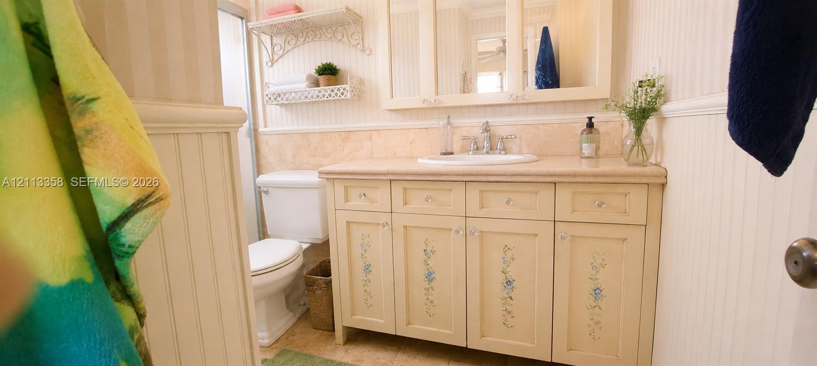 Vanity with light-toned cabinetry, inset sink with dual-handle faucet, stone-surfaced countertop, wainscoting, and wall-mounted shelving