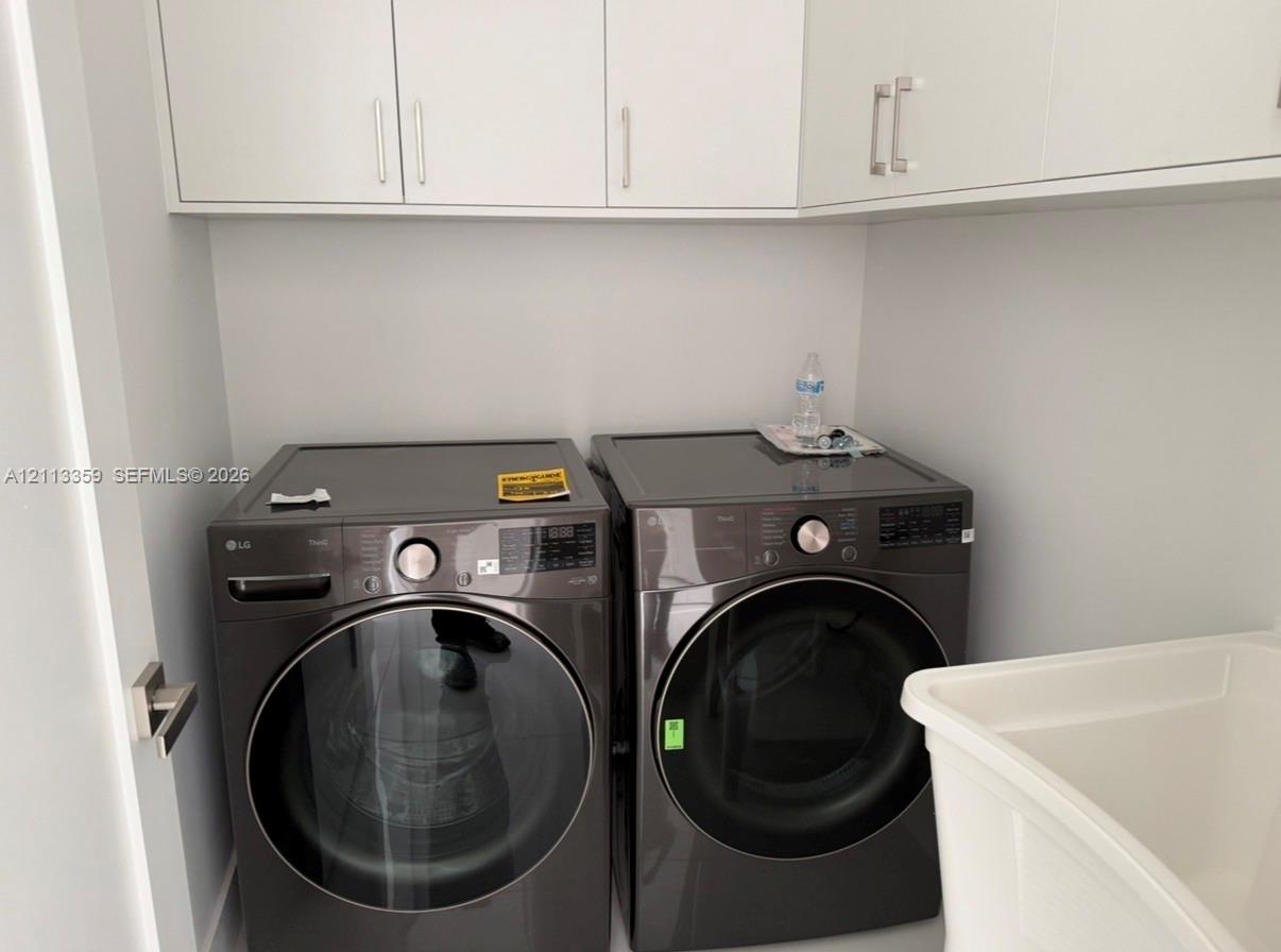 Laundry space featuring charcoal-finish front-load appliances, matte white shaker-style cabinetry with polished metal hardware, and a deep basin utility sink