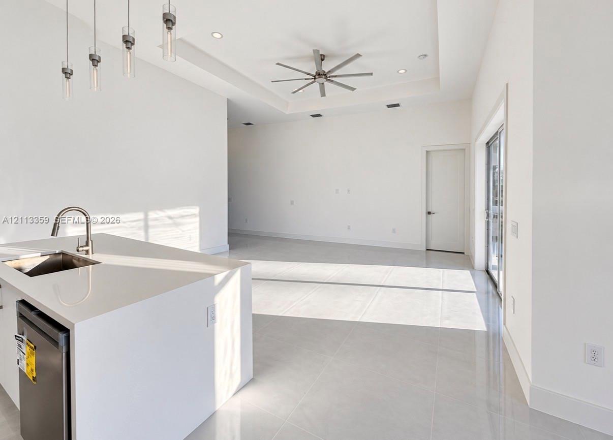 Kitchen island featuring a solid-surface countertop with an integrated undermount sink, multi-pendant lighting, recessed ceiling lighting, a tray ceiling with a multi-blade ceiling fan, and tile flooring