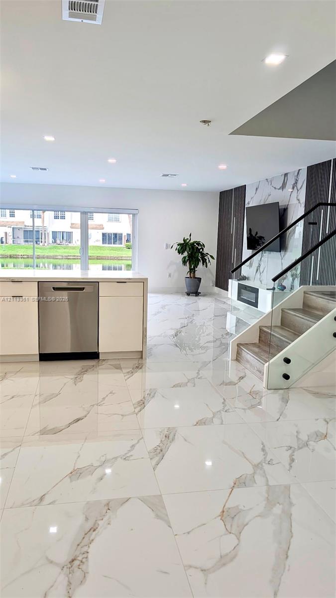 Large-format stone flooring featuring a veined pattern, white flat-panel cabinetry, stainless steel dishwasher, glass-panel staircase railing, and built-in electric fireplace with vertical accent lighting