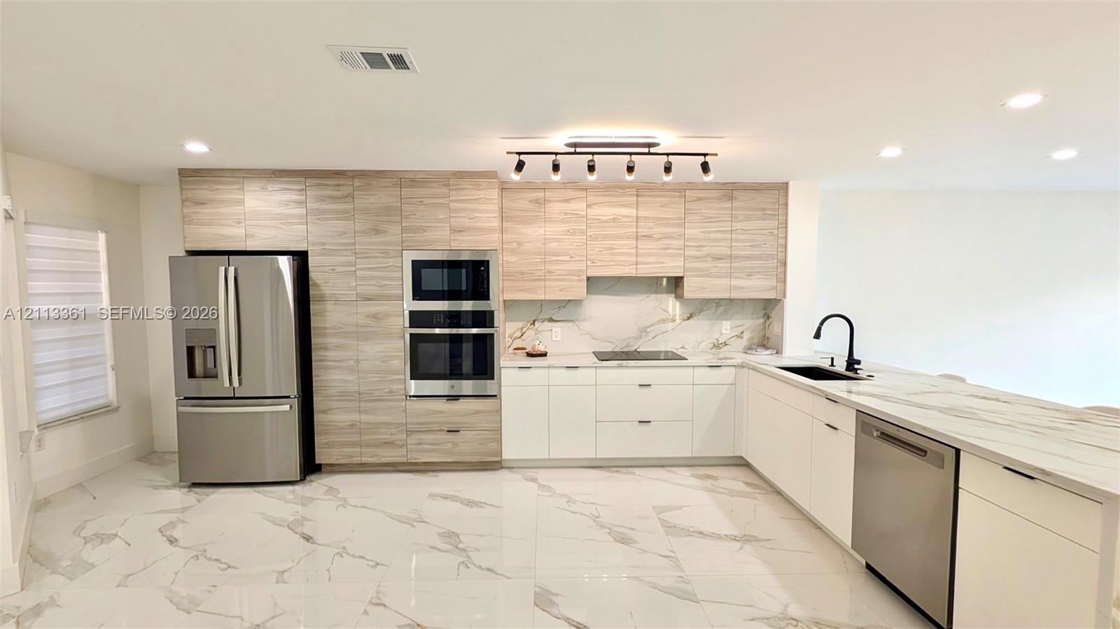 Kitchen featuring two-tone cabinetry with wood-finish upper cabinets, white lower cabinetry, integrated stainless steel appliances, stone-finish tile flooring, and multi-bulb track lighting