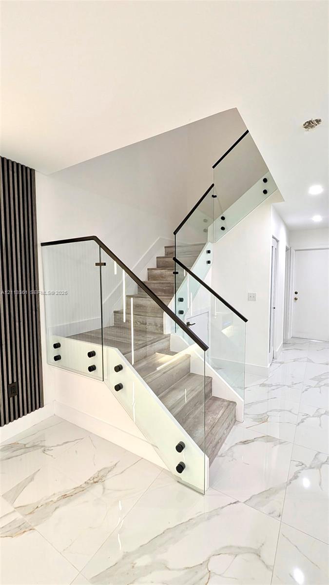 Large-format polished tile flooring, slat-wall accent paneling with integrated lighting, stone-finish fireplace surround, quartz-topped island with slat detailing, and glass-paneled staircase railing