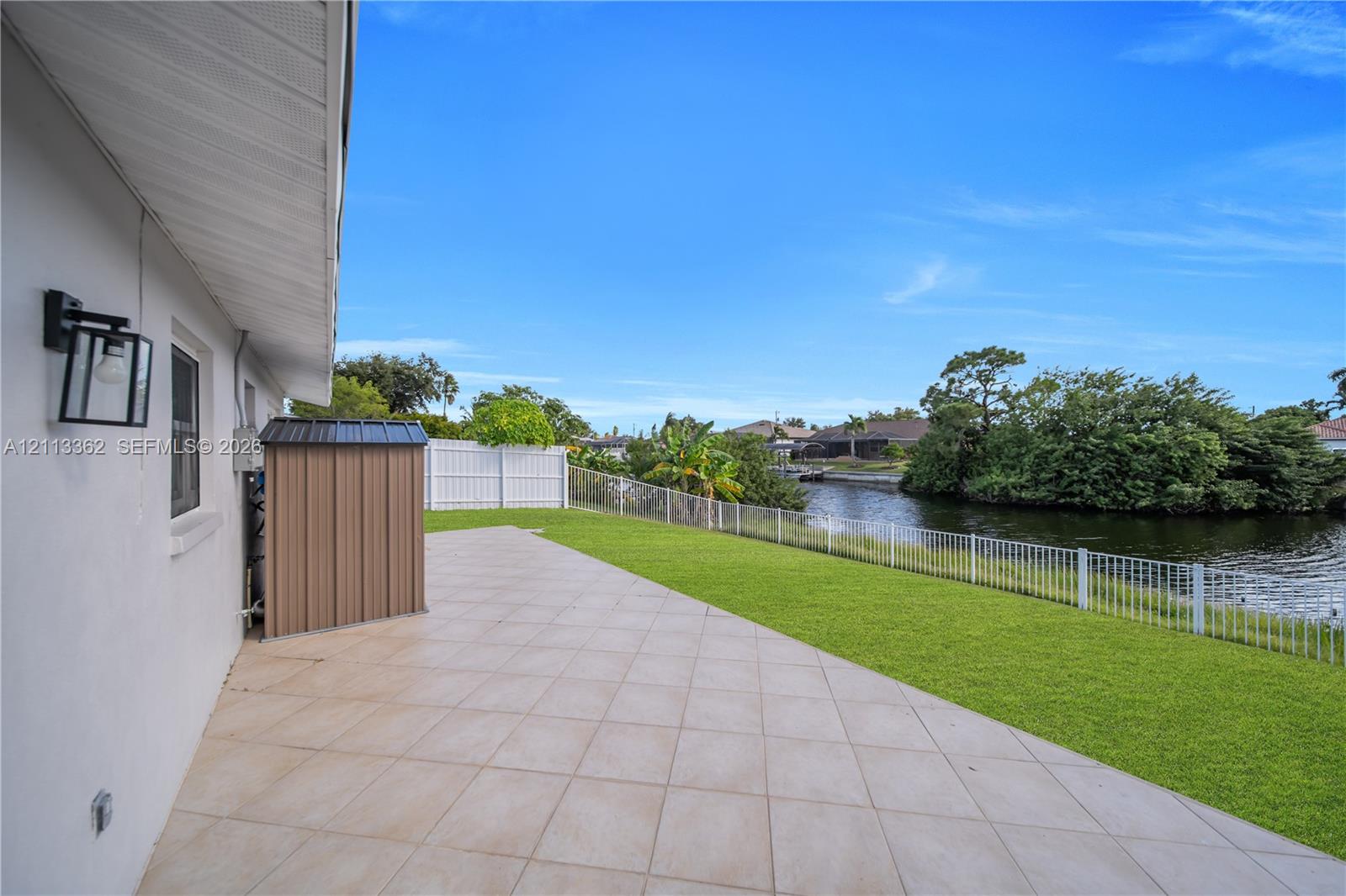 Waterfront backyard featuring a tiled patio, manicured lawn, perimeter fencing, and exterior wall-mounted lighting