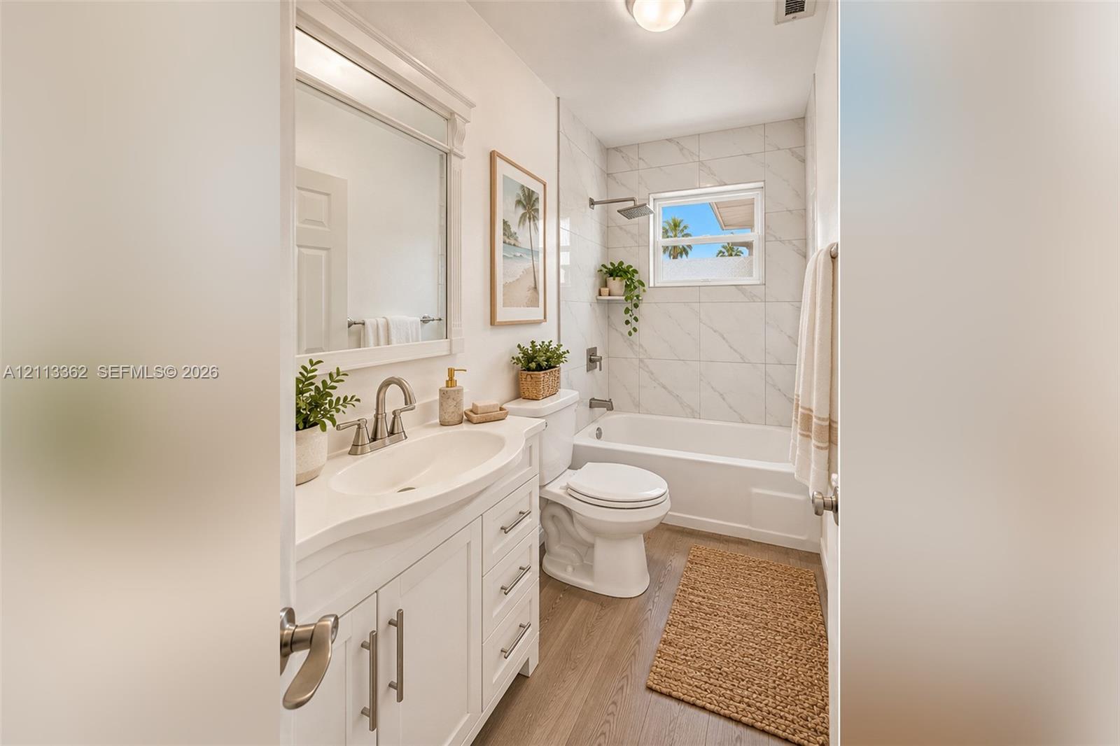 White vanity with integrated basin, polished metal hardware, framed mirror, light-toned wood-finish flooring, and tub-shower combination with floor-to-ceiling tile surround