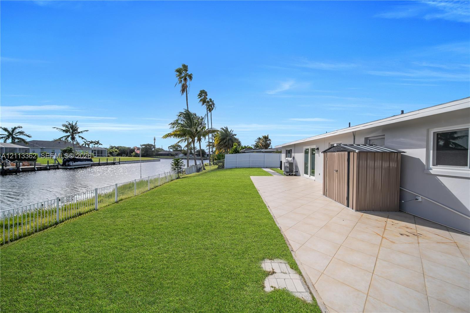Waterfront backyard featuring a manicured lawn, tiled patio, and metal perimeter fencing