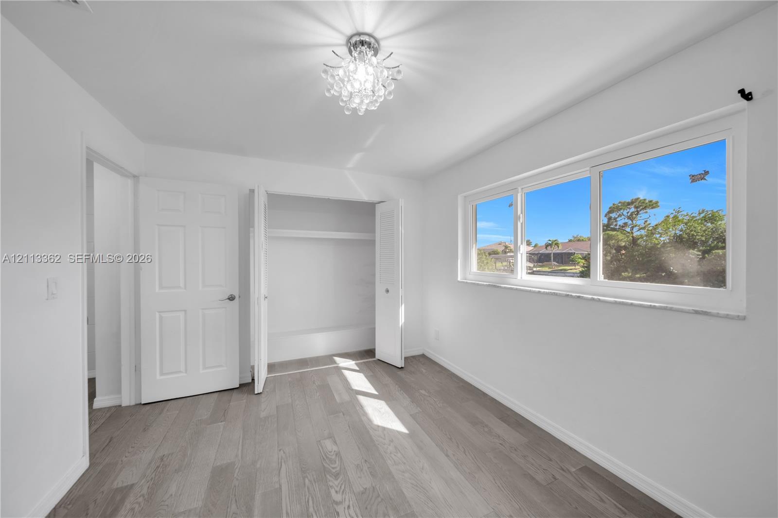 Neutral-toned wood-finish flooring, crystal-style flush mount ceiling light, wide horizontal window with stone-finish sill, and double-door closet with built-in shelving