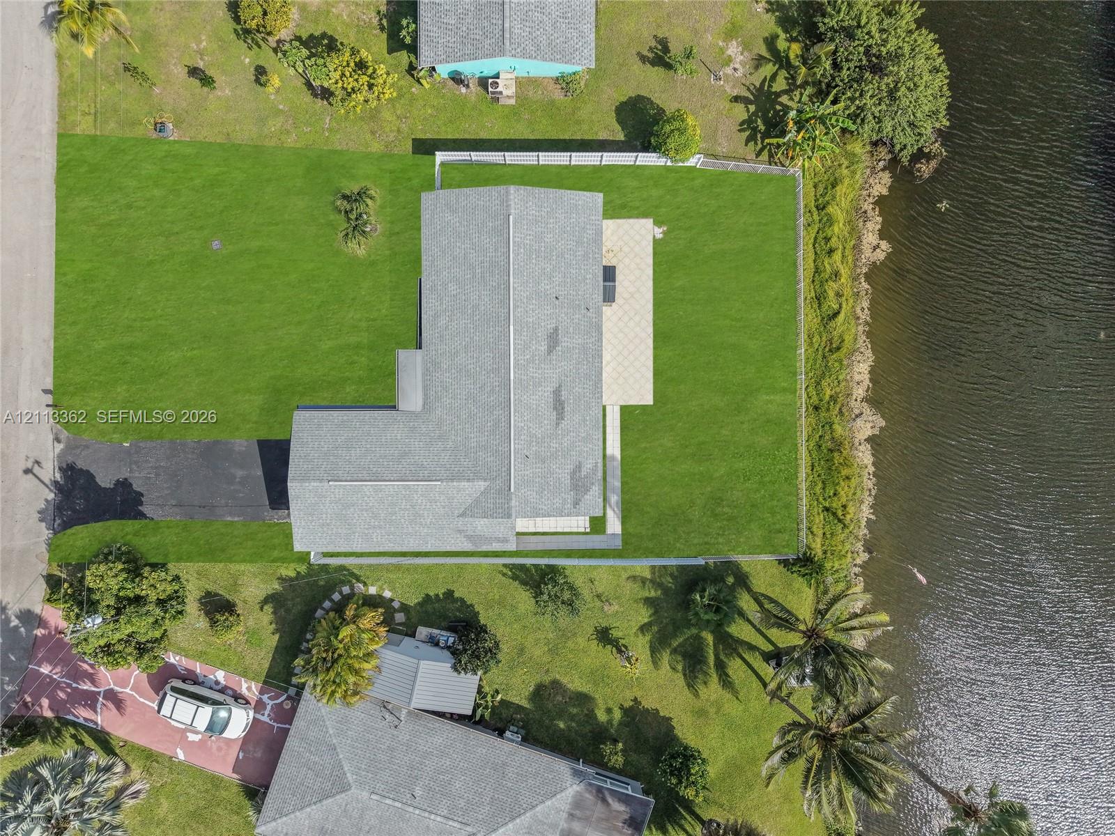 Aerial perspective featuring a waterfront lot, expansive lawn, asphalt driveway, and paved rear patio area