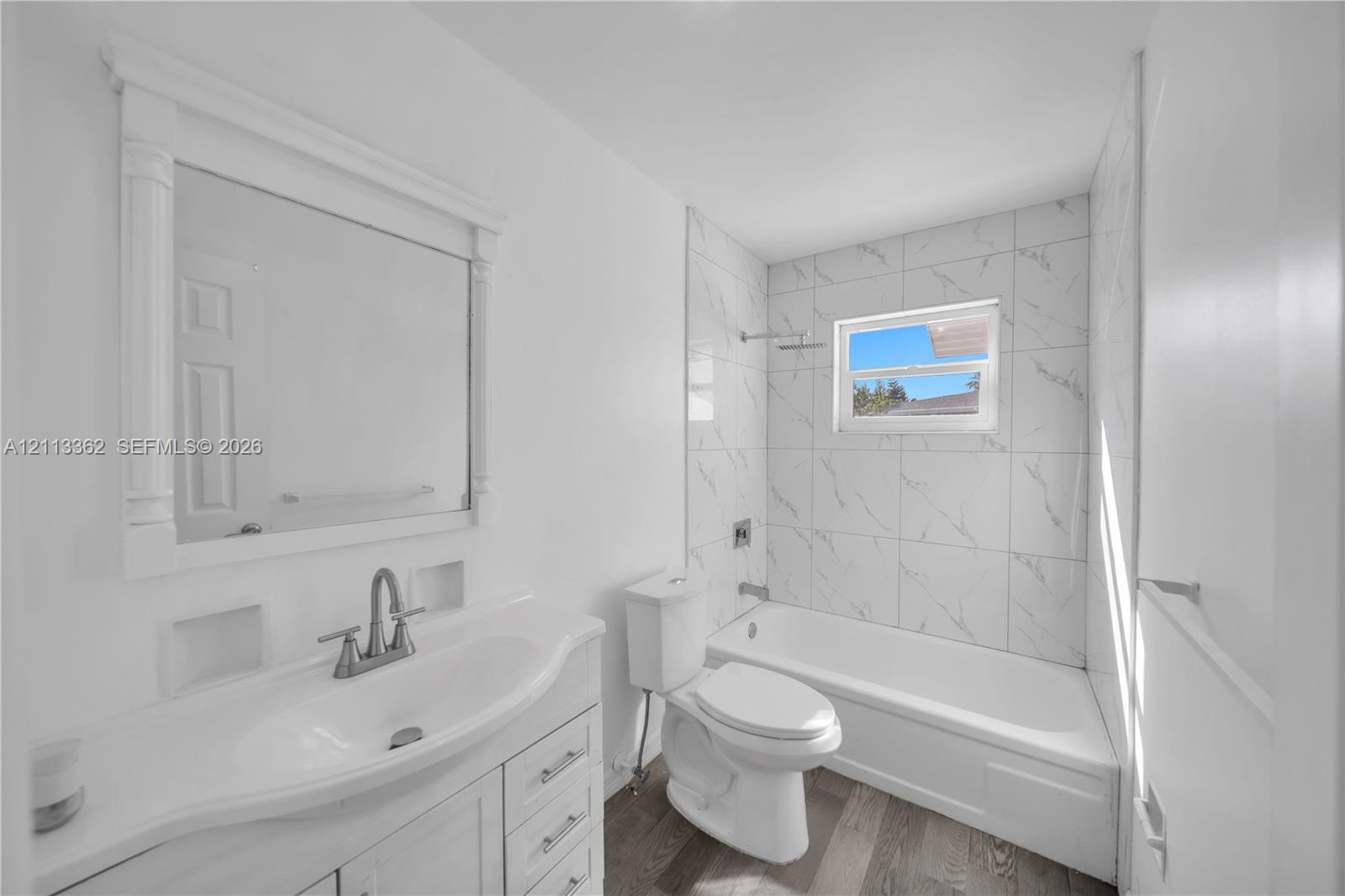 Bathroom featuring a white vanity with an integrated sink, a framed wall mirror, a bathtub with full-height stone-patterned tile surround, and wood-finish flooring