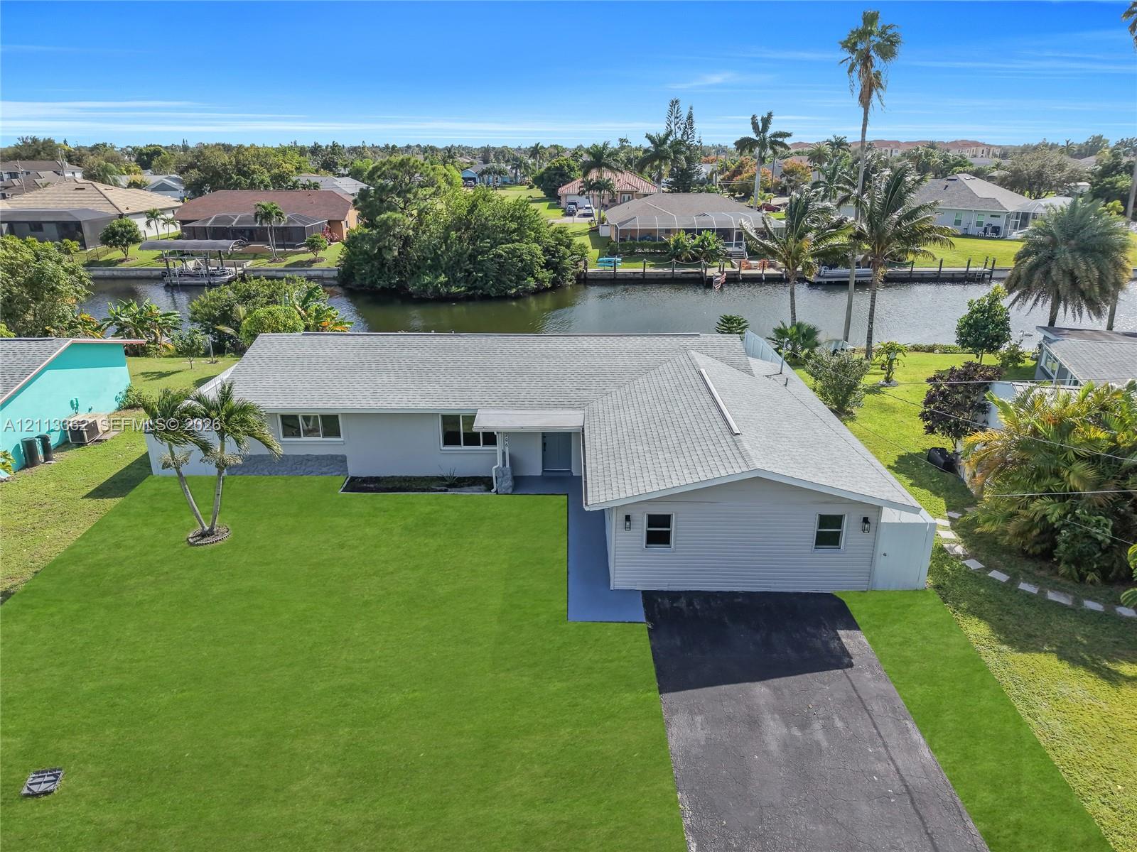 Single-story residence featuring a light-toned exterior, asphalt-shingled roof, paved driveway, expansive lawn, and canal frontage Single-story residence featuring a light-toned exterior, asphalt-shingled roof, paved driveway, expansive lawn, and canal frontage