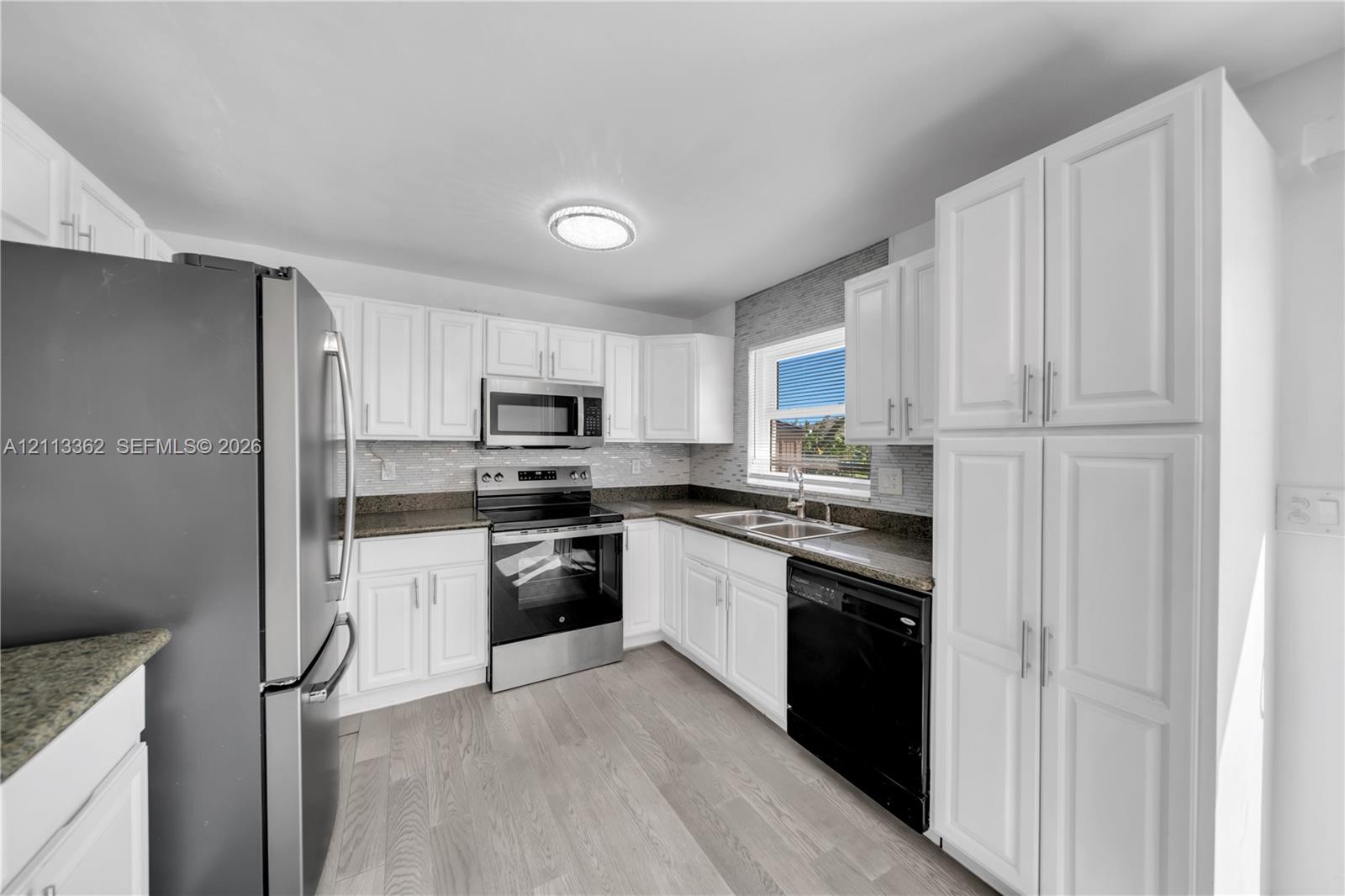 Kitchen featuring white shaker-style cabinetry, stone-finish countertops, tile backsplash, stainless steel appliances, and wood-finish flooring Kitchen featuring white shaker-style cabinetry, stone-finish countertops, tile backsplash, stainless steel appliances, and wood-finish flooring