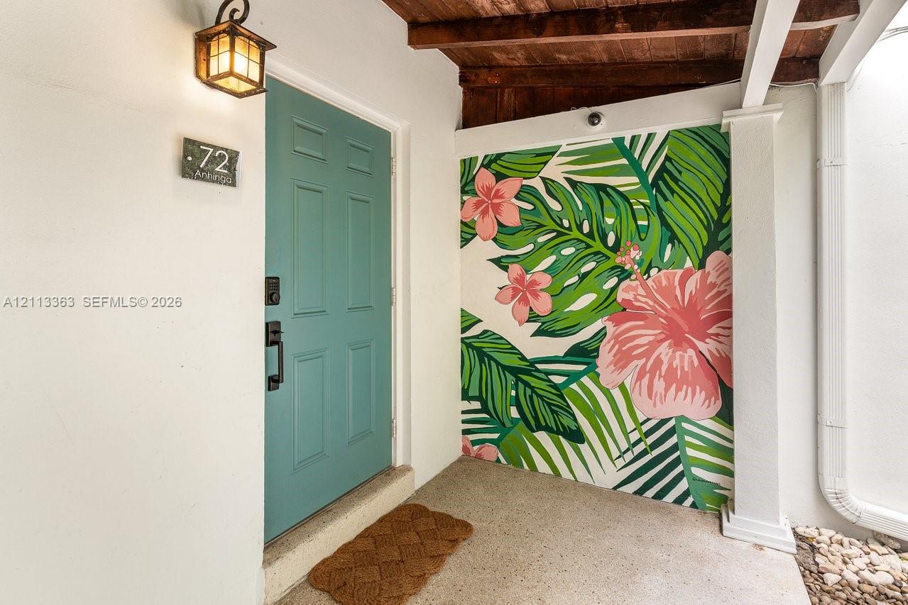 Entryway featuring a solid teal six-panel door, custom floral wall mural, exposed wood ceiling beams, and a wall-mounted lantern-style fixture Entryway featuring a solid teal six-panel door, custom floral wall mural, exposed wood ceiling beams, and a wall-mounted lantern-style fixture