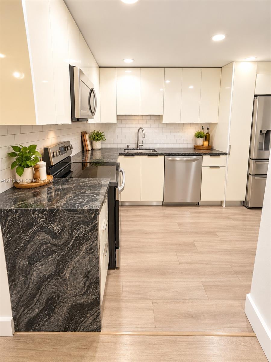 Kitchen featuring flat-panel high-gloss cabinetry, dark stone countertops with waterfall edge, white subway tile backsplash, stainless steel appliances, and wood-finish flooring