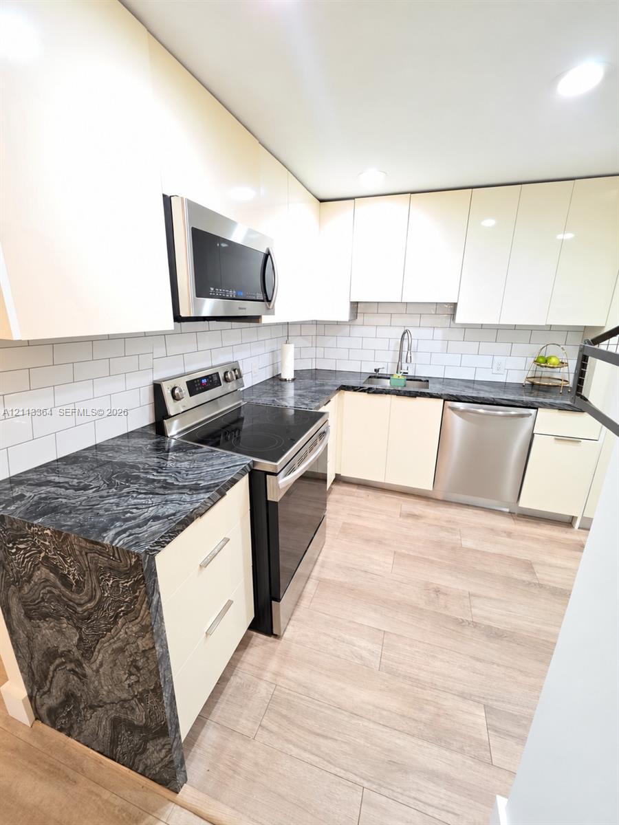 Kitchen featuring high-gloss cabinetry, stone countertops with waterfall edge, stainless steel appliances, subway tile backsplash, and wood-finish tile flooring