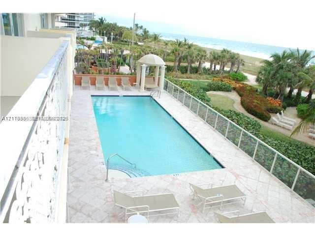 Rectangular swimming pool featuring a tile deck, glass panel perimeter railing, and a columned cabana structure, situated against a sandy coastline backdrop