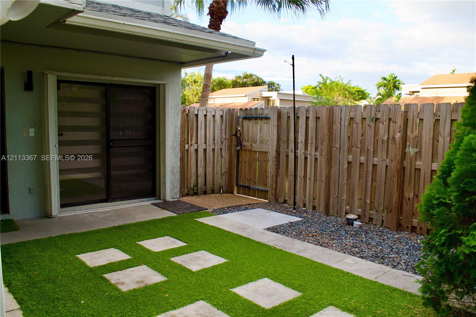 Private outdoor patio area featuring artificial turf, concrete pavers, a gravel landscape border, and a wood privacy fence with a gated entry
