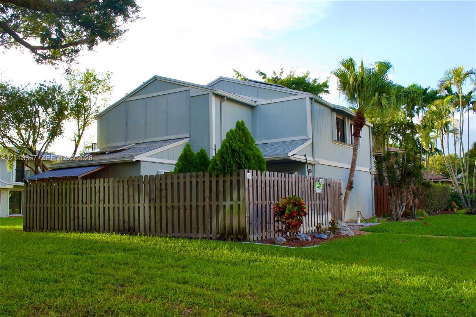 Two-story exterior featuring vertical siding, a shingled roof, a wooden privacy fence, and mature palm trees