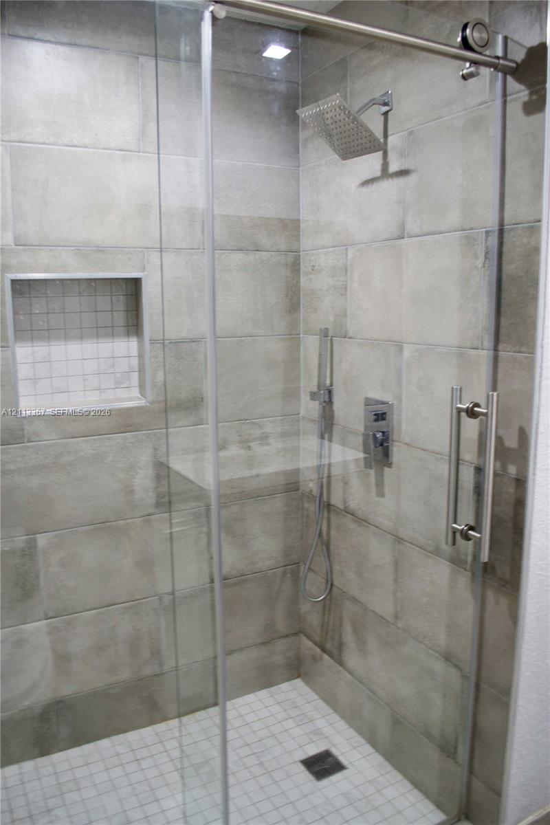 Walk-in shower featuring large-format neutral tile walls, a sliding glass door with stainless steel hardware, a rainfall showerhead, a handheld sprayer, and a built-in recessed niche