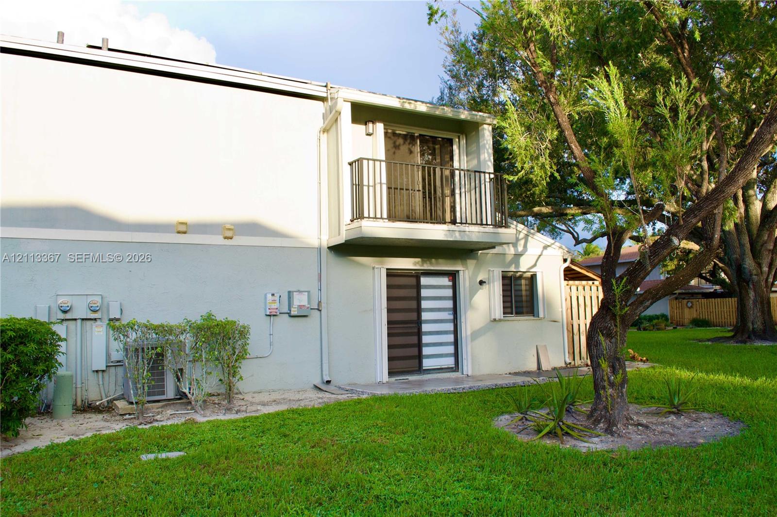 Exterior featuring a second-story balcony with metal railing, sliding glass doors, and a ground-level grass yard with mature trees