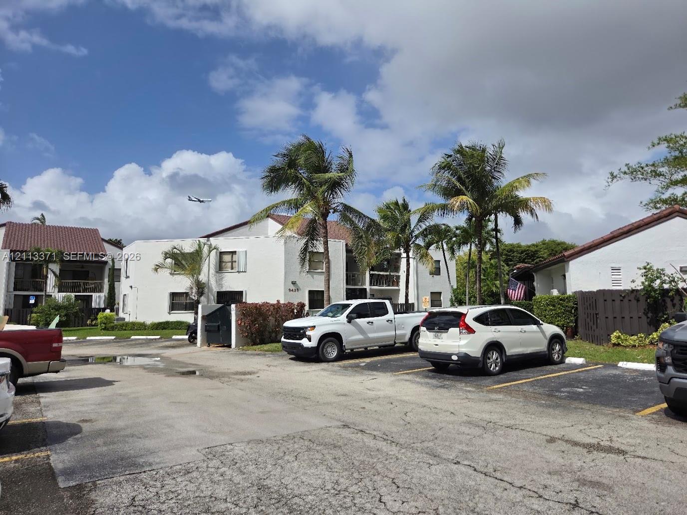 Multi-story residential building featuring white stucco exterior, tile roofing, balcony railings, and mature palm trees