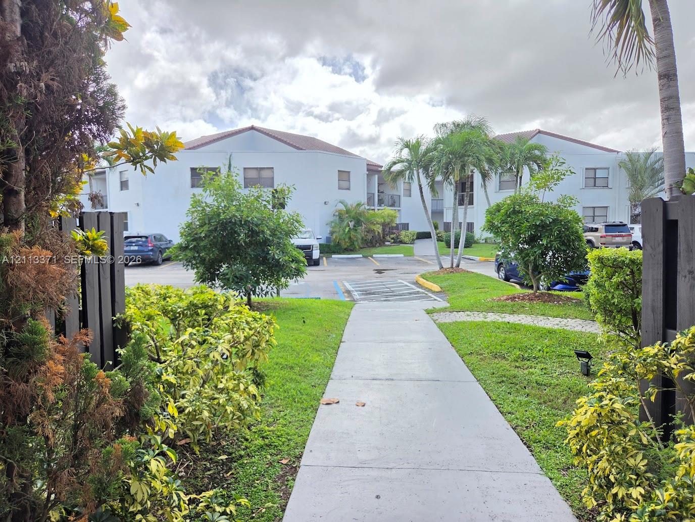 Multi-story building exterior with a light-colored stucco facade and tile roof, concrete walkway, mature palm trees, and landscaped grounds