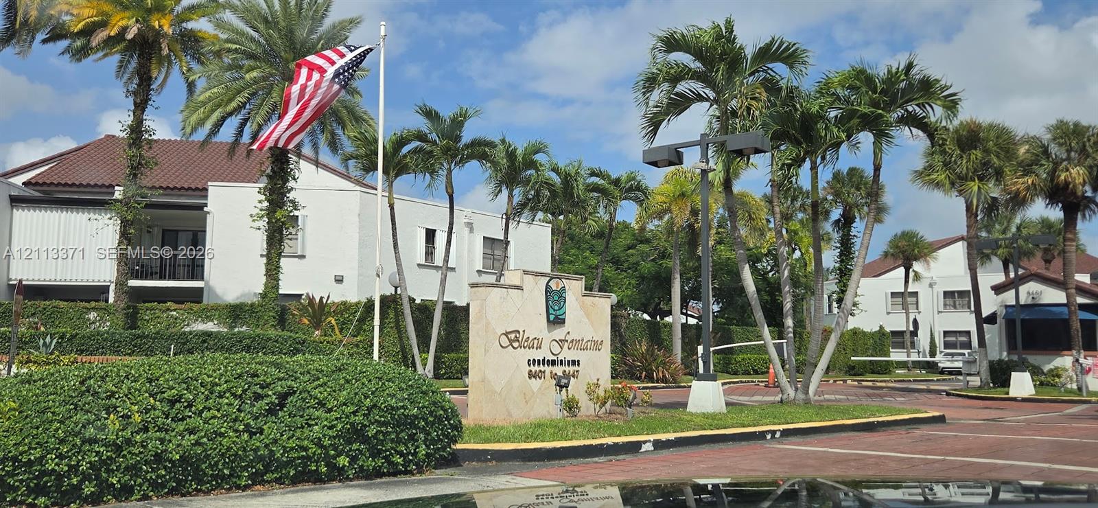 Stone community monument sign featuring mature palm tree landscaping, paved entryway, and multi-level stucco structures