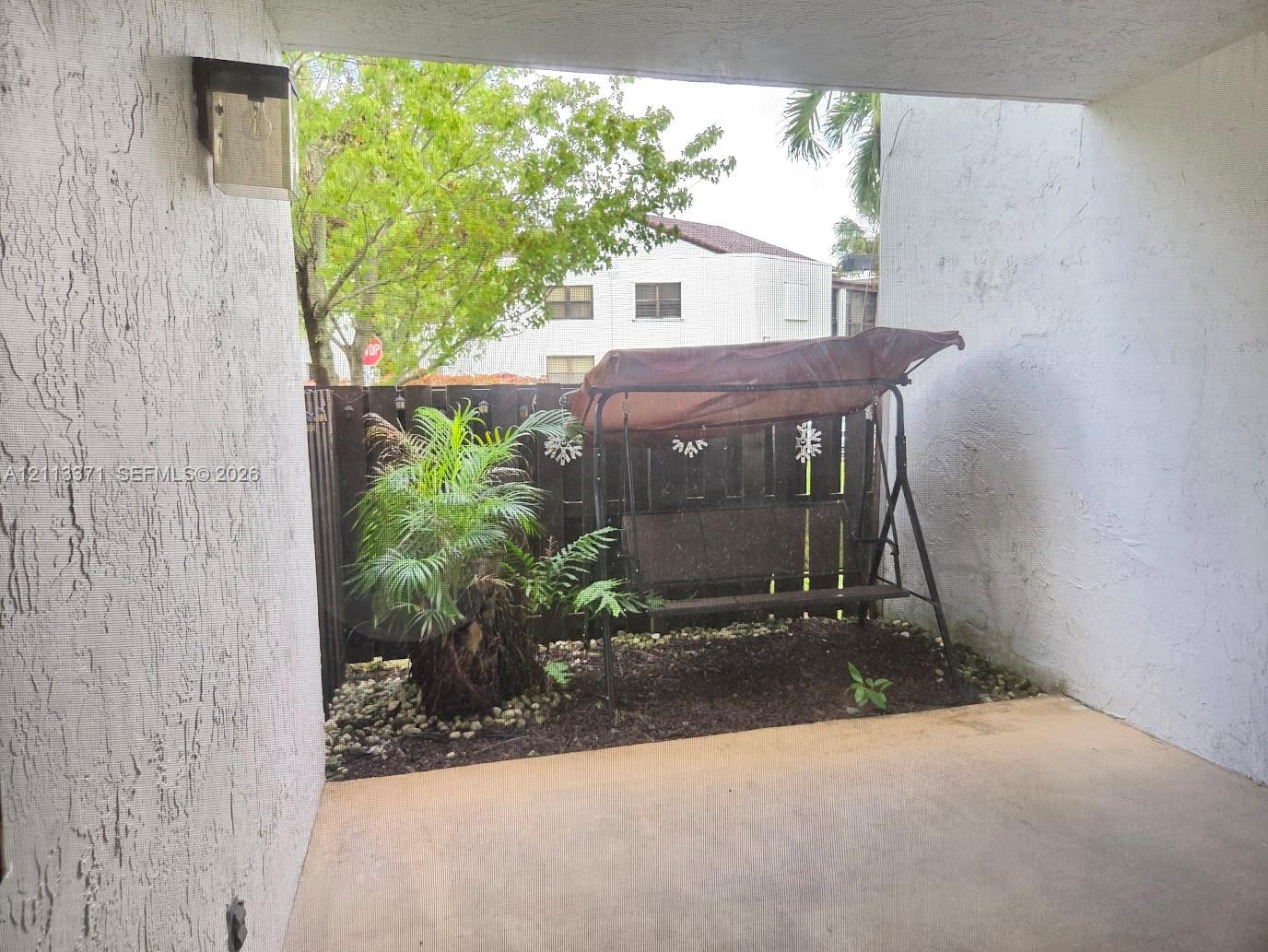 Covered concrete patio featuring stucco walls, wall-mounted exterior lighting, and a fenced privacy area with garden landscaping