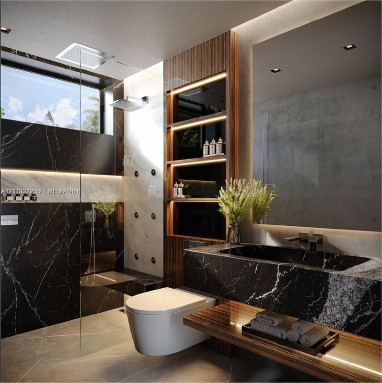 Black stone vanity with an integrated basin, wall-mounted commode, glass-enclosed shower featuring body sprays, wood-finish built-in shelving with integrated lighting, and a large rectangular mirror with perimeter illumination