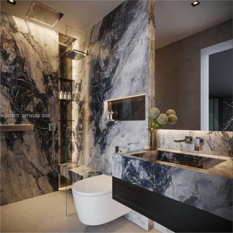 Bathroom featuring large-format stone wall cladding, floating vanity with integrated stone sink, wall-mounted faucet, recessed shelf with accent lighting, and a glass-enclosed shower with a rainfall showerhead