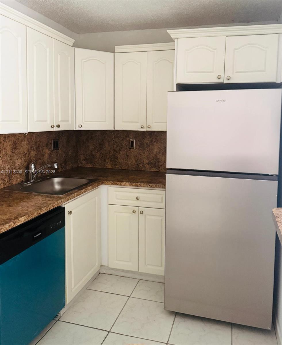 Kitchen featuring white shaker-style cabinetry, a stainless steel refrigerator, patterned laminate countertops, a stainless steel sink with a high-arch faucet, and ceramic tile flooring